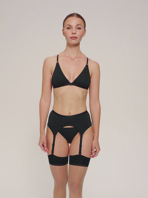 A model wearing vintage-style beige sheer stockings with black trim, paired with a black suspender belt and black bikini lingerie set.