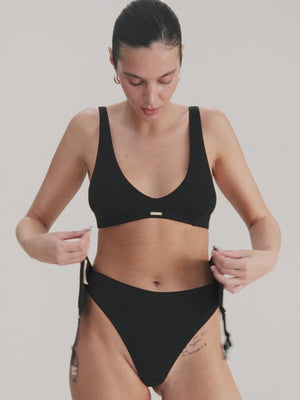 Woman wearing a black lingerie set and putting on a suspender belt and sheer stockings on a light background